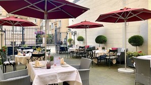 Un espace extérieur aménagé pour les repas, avec des tables et des chaises, des parasols rouges et un bâtiment en arrière-plan.
