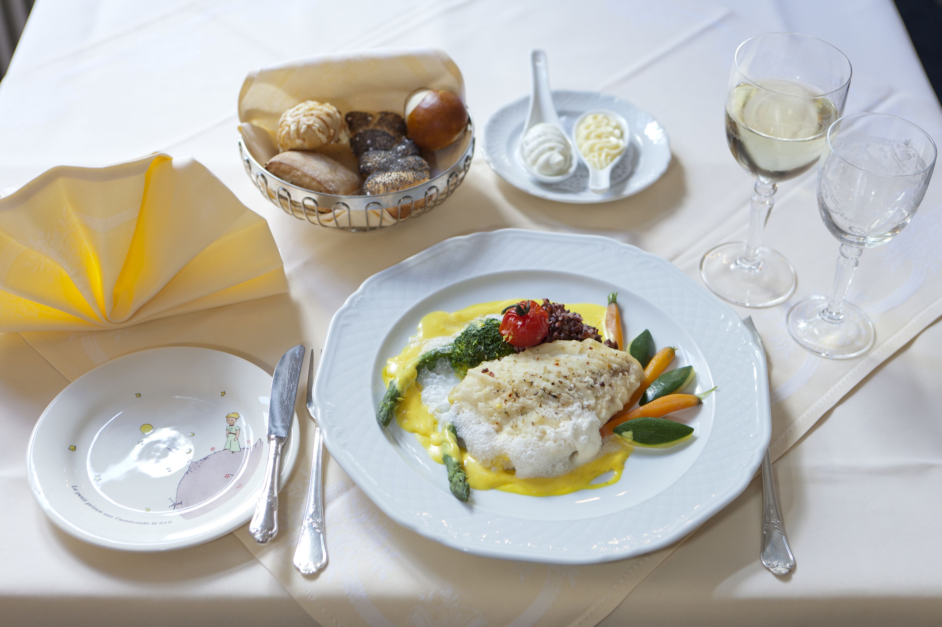 Una mesa dispuesta con un plato de pescado, verduras y salsa, un bol de pan, una tabla con mantequilla y una cuchara, y dos copas de vino blanco.