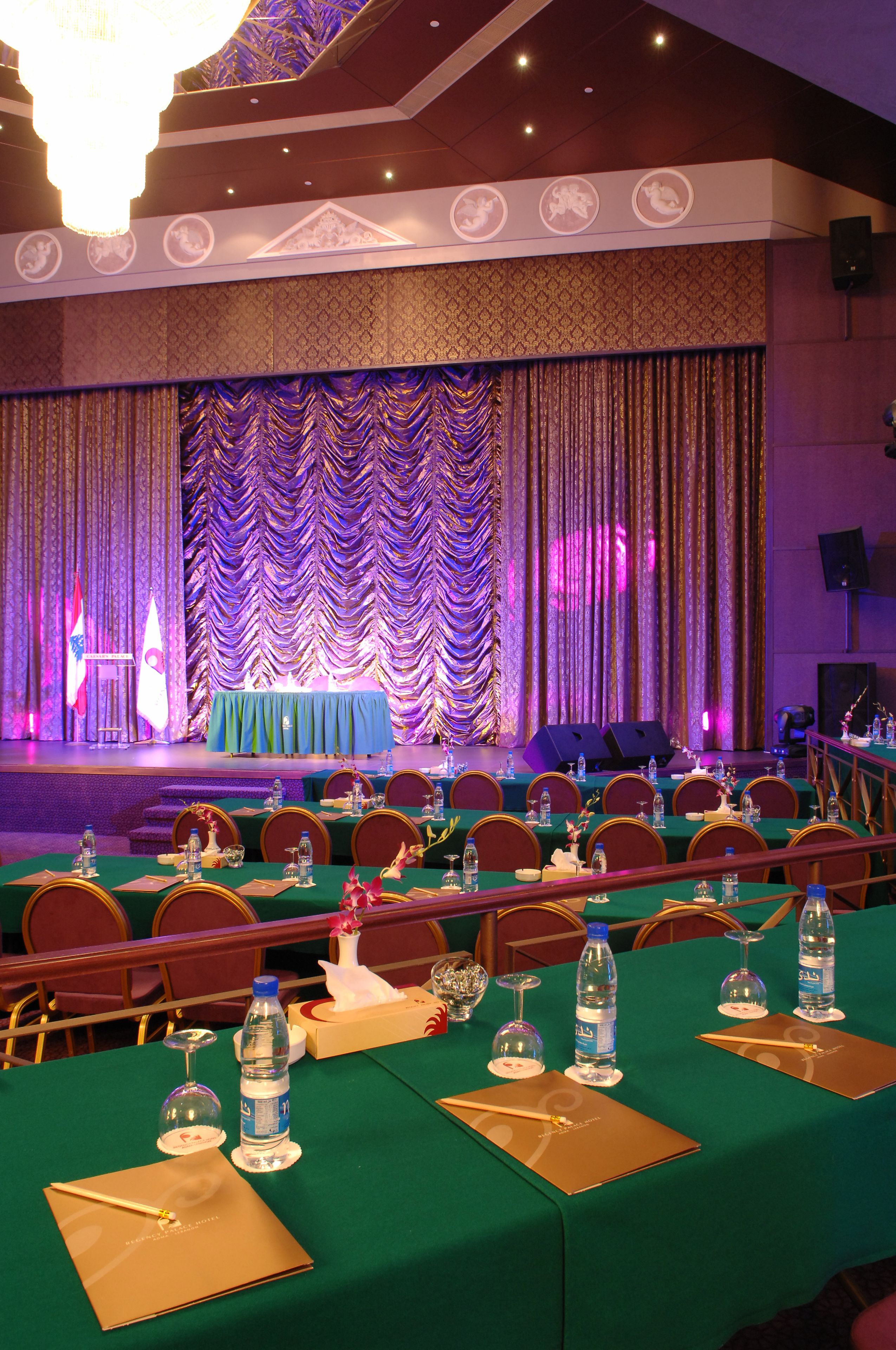A conference room with tables set for a meeting, a stage with curtains, and a chandelier.
