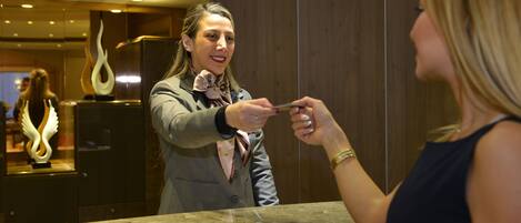 Una persona con traje y corbata de pie frente al mostrador de recepción en el vestíbulo de un hotel, con una encimera de mármol y una escultura decorativa al fondo.