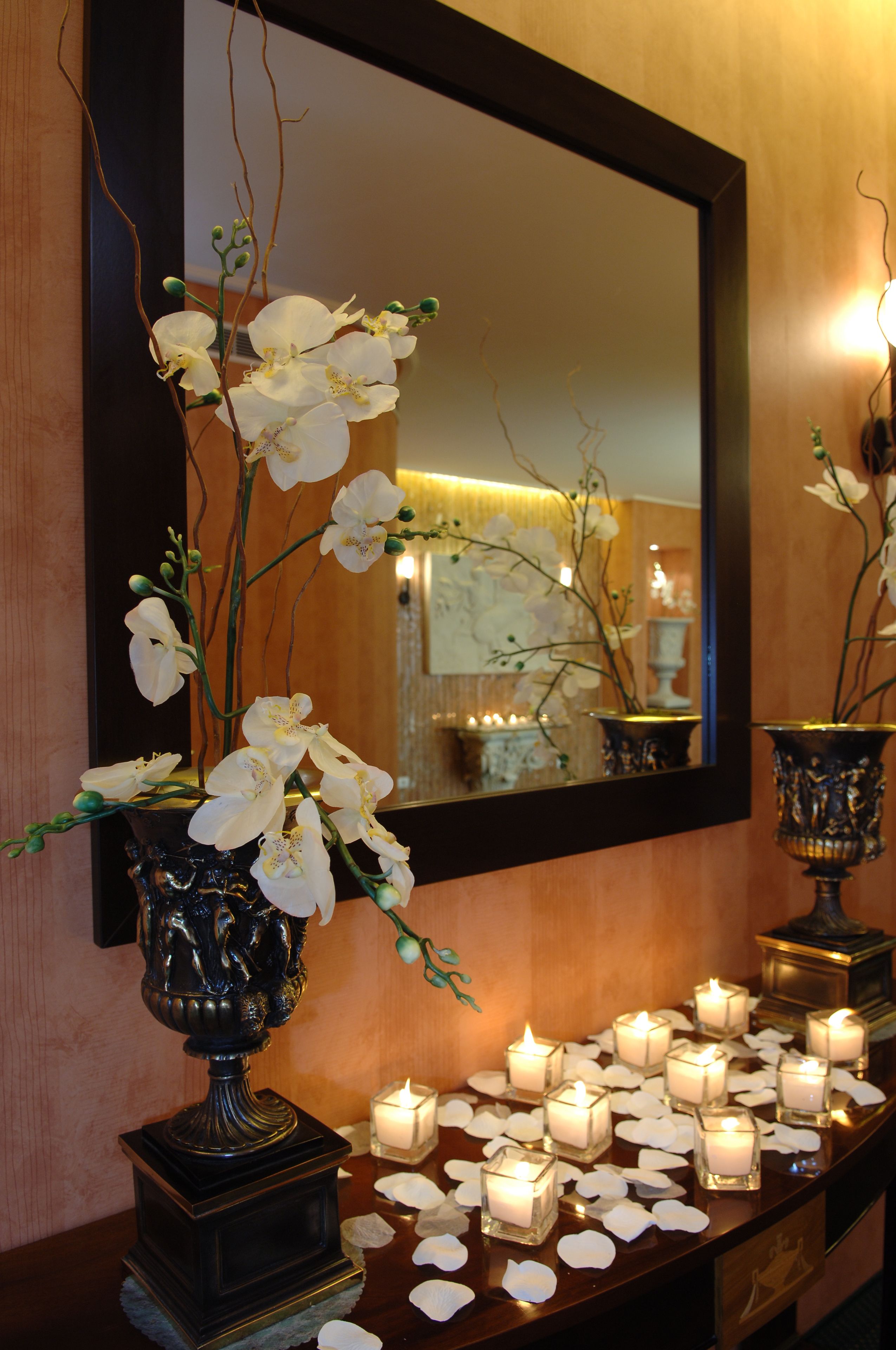 A reception area with a mirror, a vase with white flowers, and a row of lit candles on a wooden table.