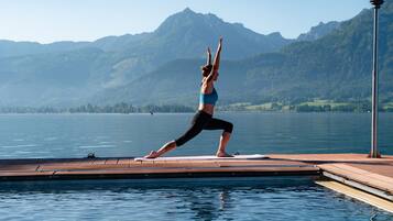 A person performing a yoga pose on a wooden dock by a lake with mountains in the background.