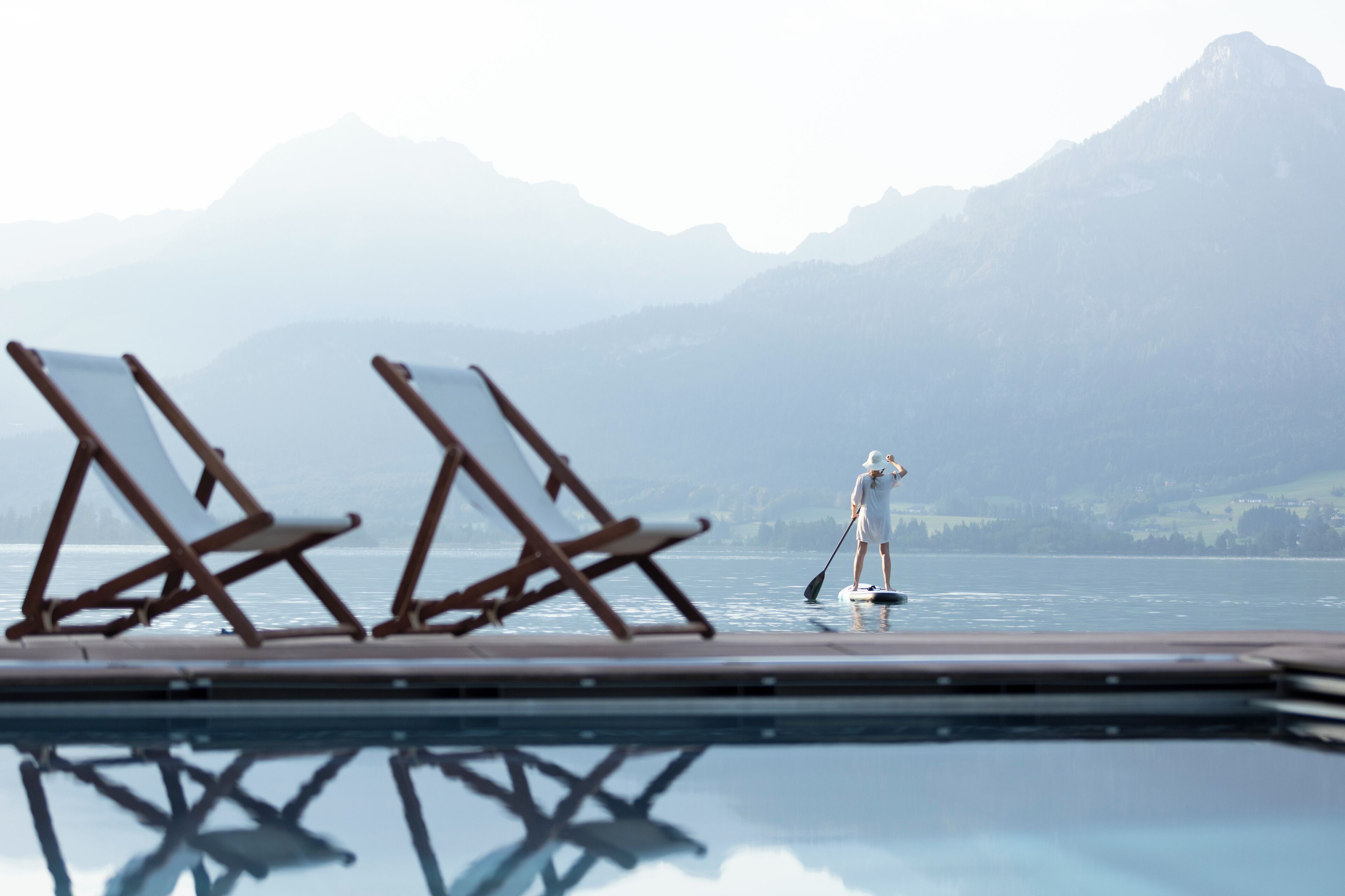 A deck with two lounge chairs facing a calm body of water, a person paddleboarding, and a mountain range in the distance.