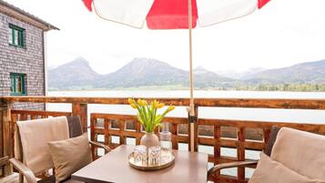 Eine Holzterrasse mit Tisch und Stühlen, einem roten und weißen Sonnenschirm und Blick auf Berge und Wasser.