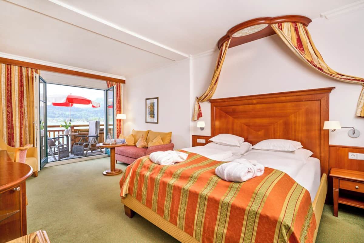 A hotel room with a large bed, a wooden headboard, a balcony with a red umbrella, and a view of the landscape.