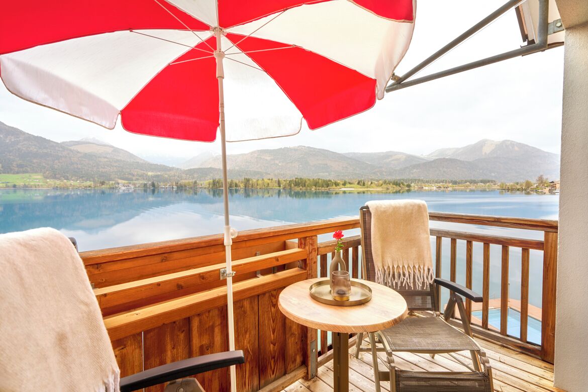 A wooden deck with a table and chairs, a red and white umbrella, and a view of a lake and mountains.