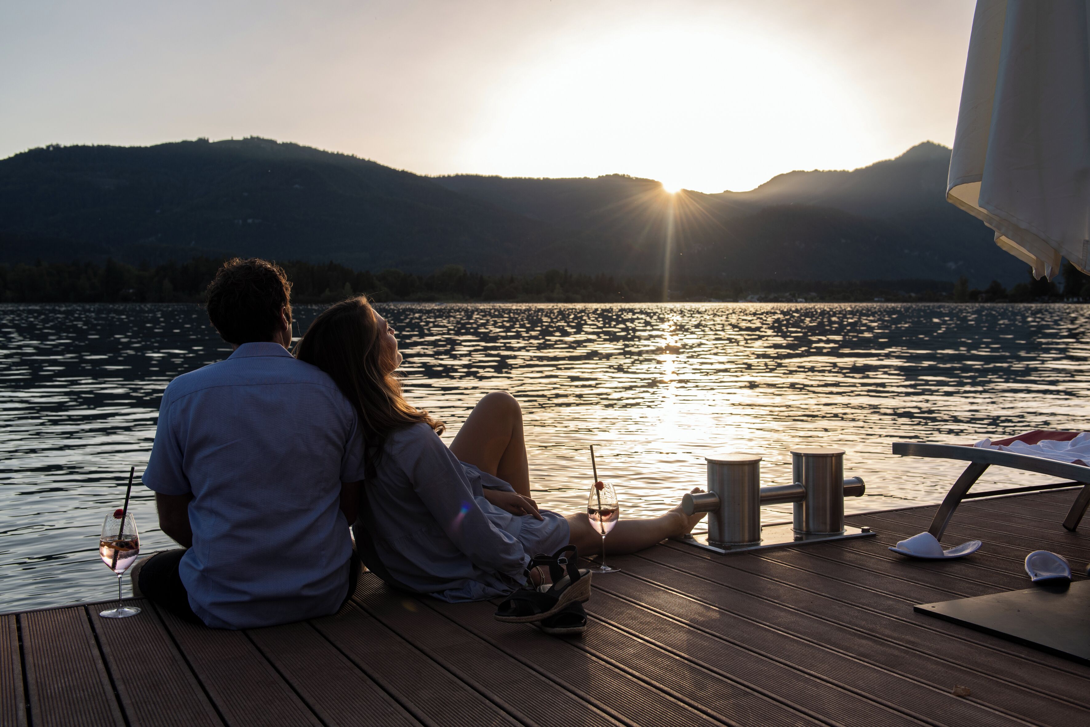 A couple sitting on a wooden deck overlooking a body of water with mountains in the background.