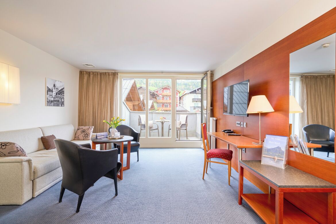A hotel room with a sofa, a dining table with chairs, a desk with a chair, a TV, and a large window with curtains.