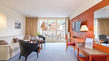 A hotel room with a sofa, a dining table with chairs, a desk with a chair, a TV, and a large window with curtains.