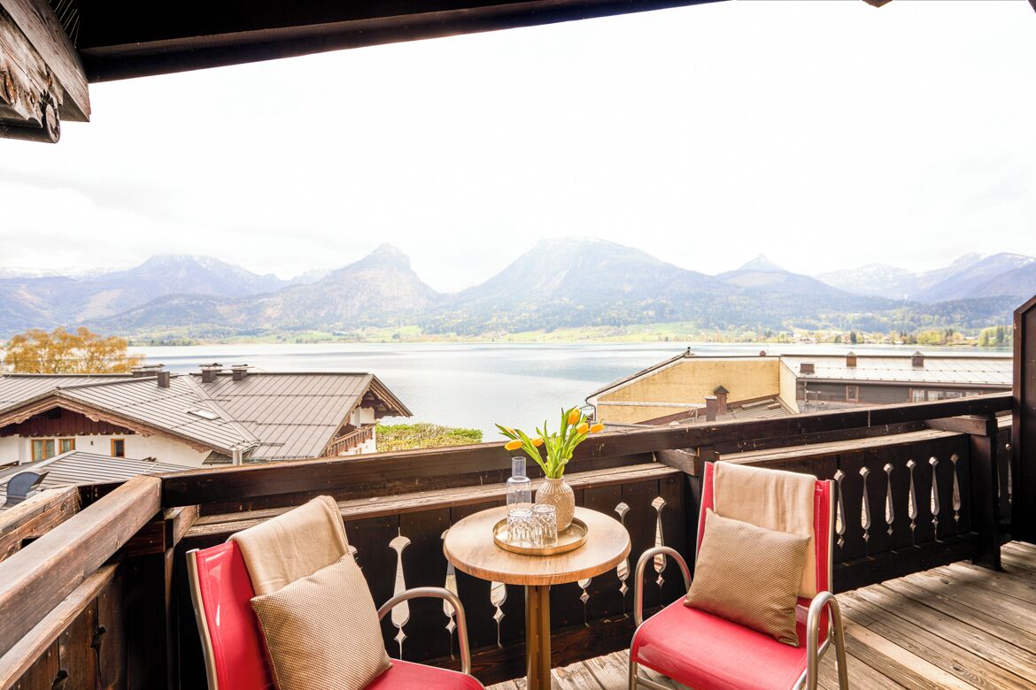 Een balkon met uitzicht op bergen en een meer, met houten meubels en een kleine tafel met een vaas.