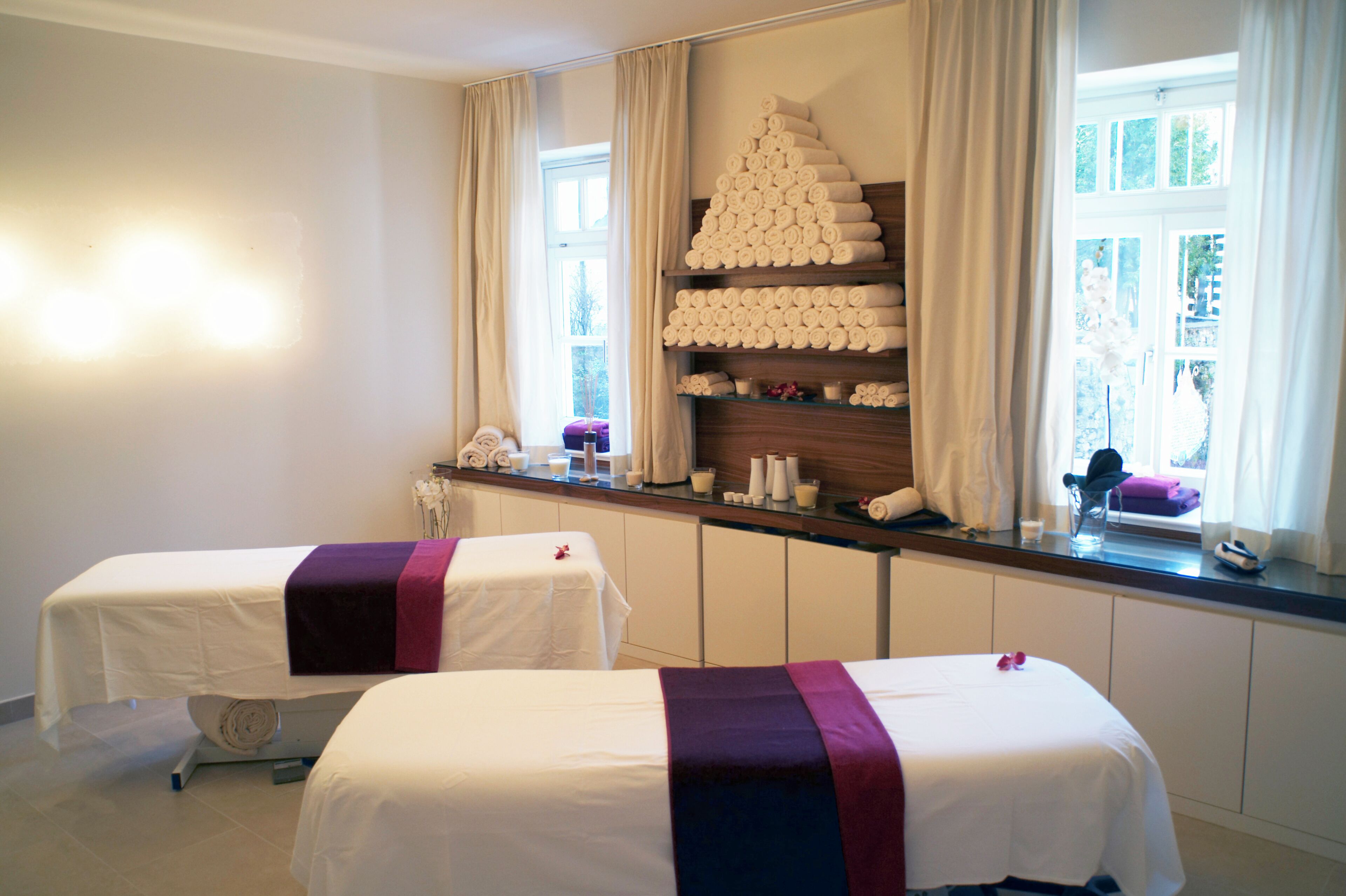A room with two beds, a window with curtains, a shelf with towels, and a small table with toiletries.