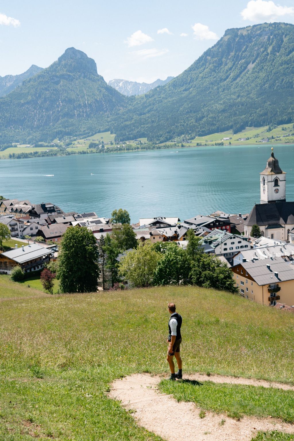 Une personne se tient sur une colline herbeuse, d’où elle a une vue sur un lac, avec des montagnes en arrière-plan et une ville où l’on aperçoit des bâtiments et le clocher d’une église.