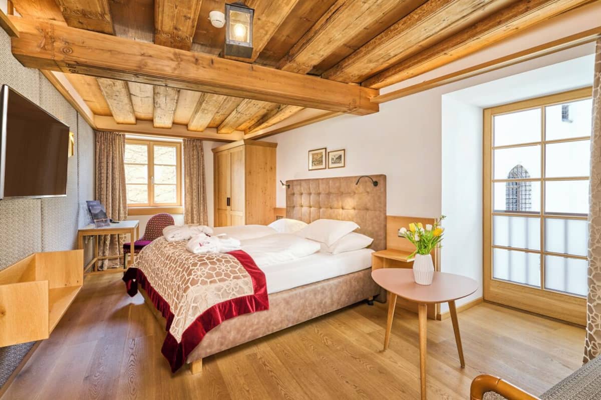 A hotel room with a large bed, wooden ceiling beams, a desk, and a window with a city view.