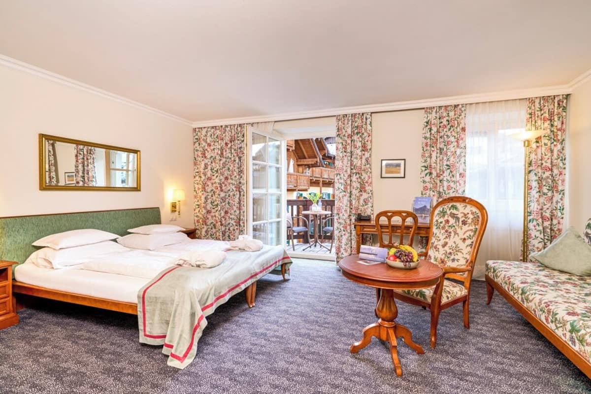 A hotel room with a bed, a dining table with chairs, a sofa, a mirror, and a view of the outside.