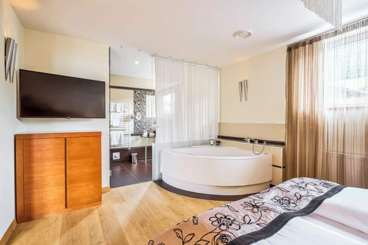 A modern hotel room with a large bed, a round bathtub, a flat-screen TV, and a wooden cabinet.