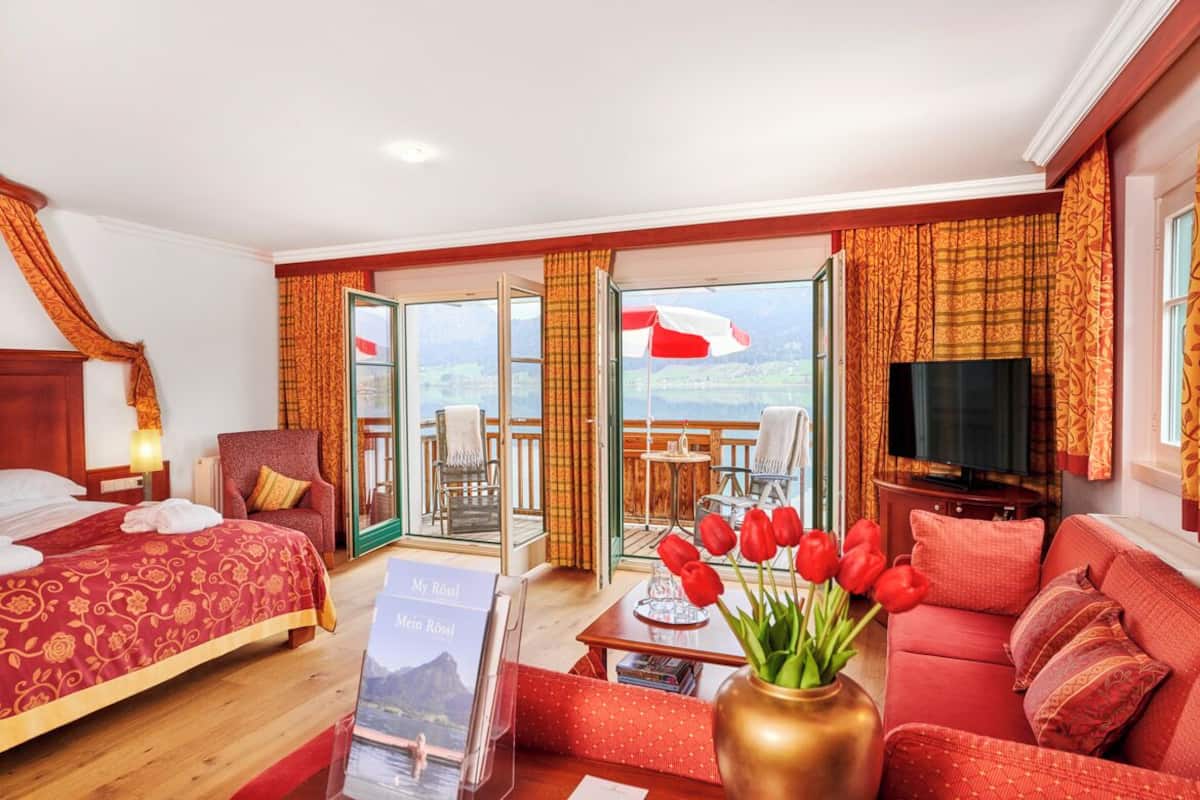 A hotel room with a bed, a sofa, a TV, a table with a vase of flowers, and a balcony with a view.