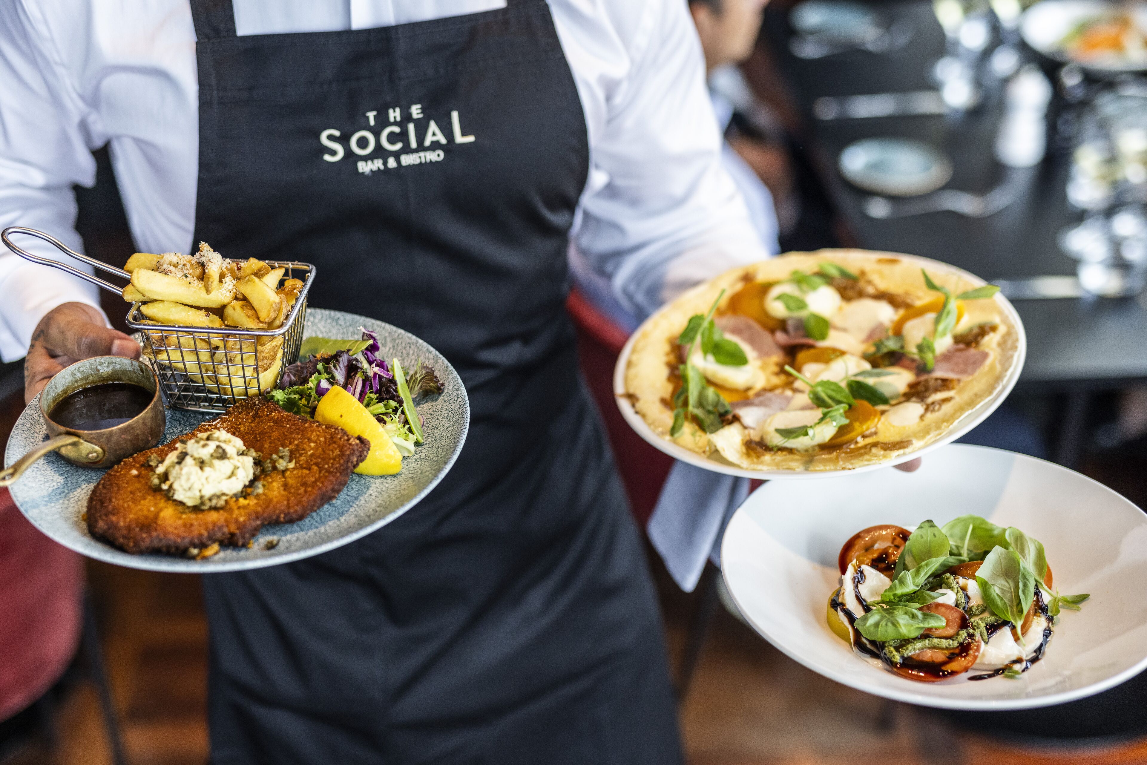 Eine Person hält einen Teller mit einem panierten Schnitzel, Pommes und Salat, und einen weiteren Teller mit einer Pizza und Salat.