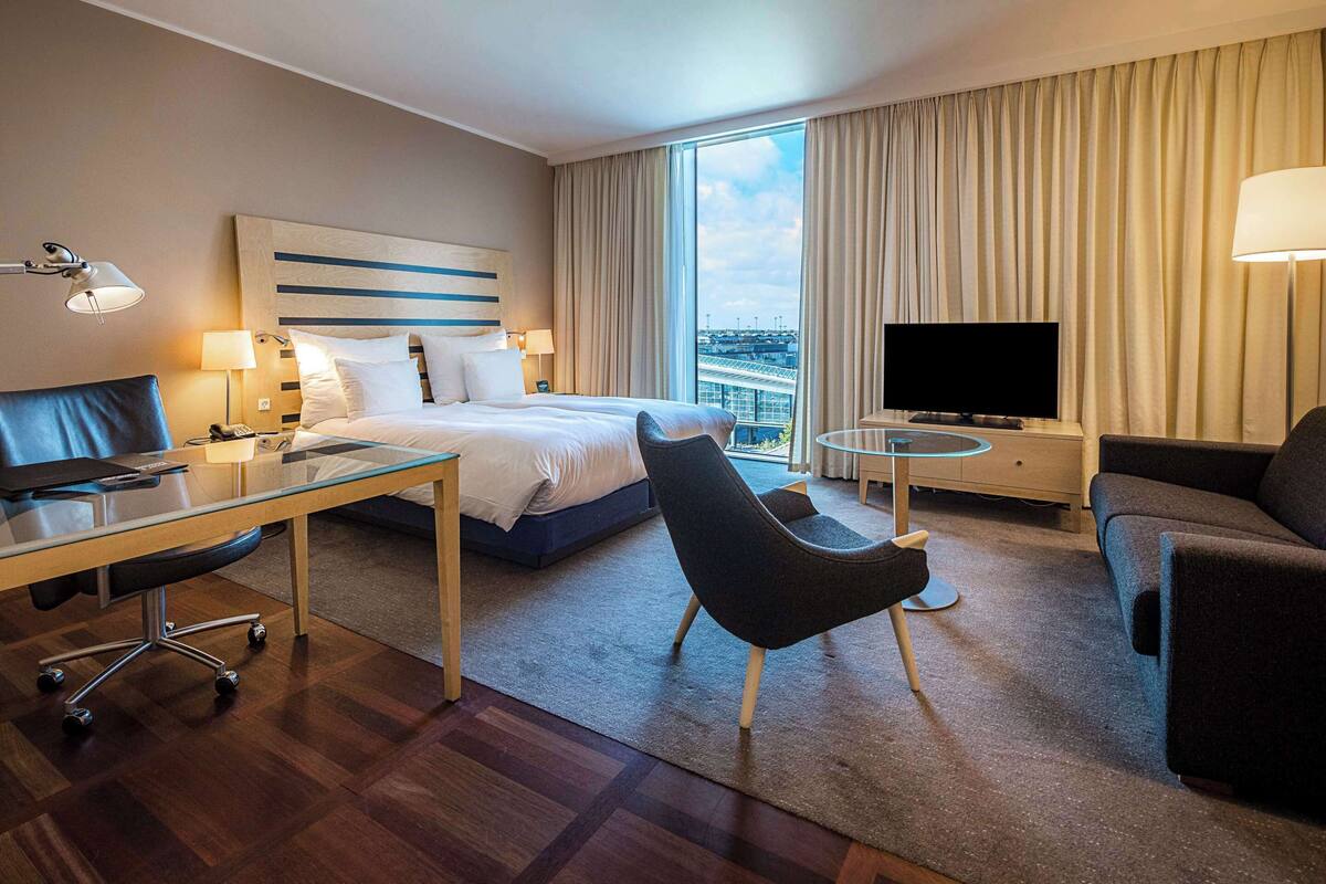 Ein Hotelzimmer mit Bett, Schreibtisch, Sessel, Sofa, Fernseher und Blick auf die Stadt.