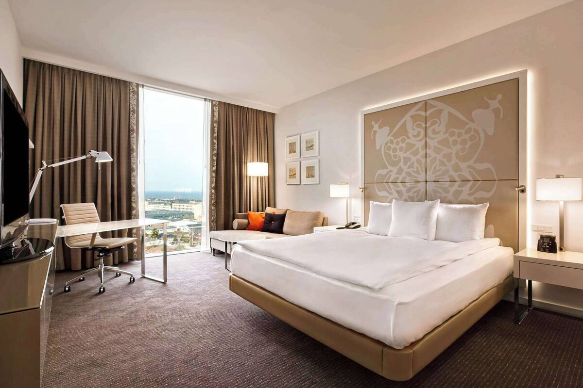 Ein Hotelzimmer mit einem großen Bett, einem Schreibtisch, einem Sessel, einer Couch und Blick auf die Stadt.