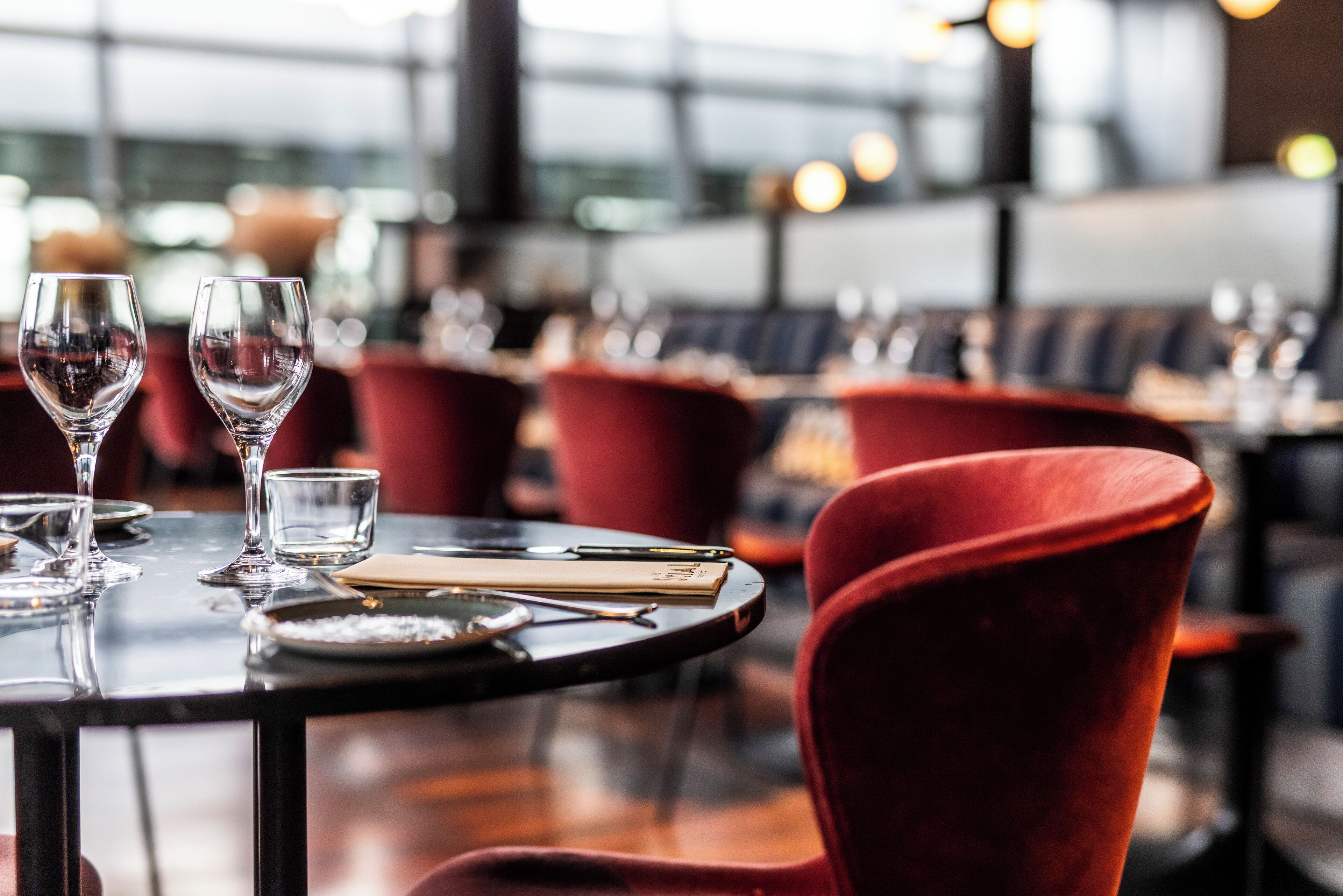 A restaurant setting with a table set for two, featuring red upholstered chairs and a glass wine glass.
