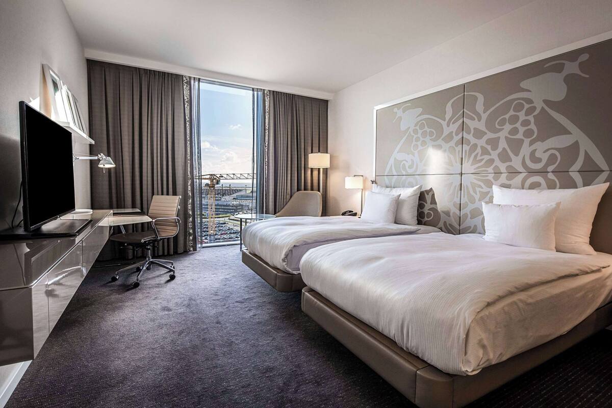 Ein Hotelzimmer mit einem großen Bett, einem Schreibtisch mit Fernseher, einem Sessel und Blick auf die Stadt.