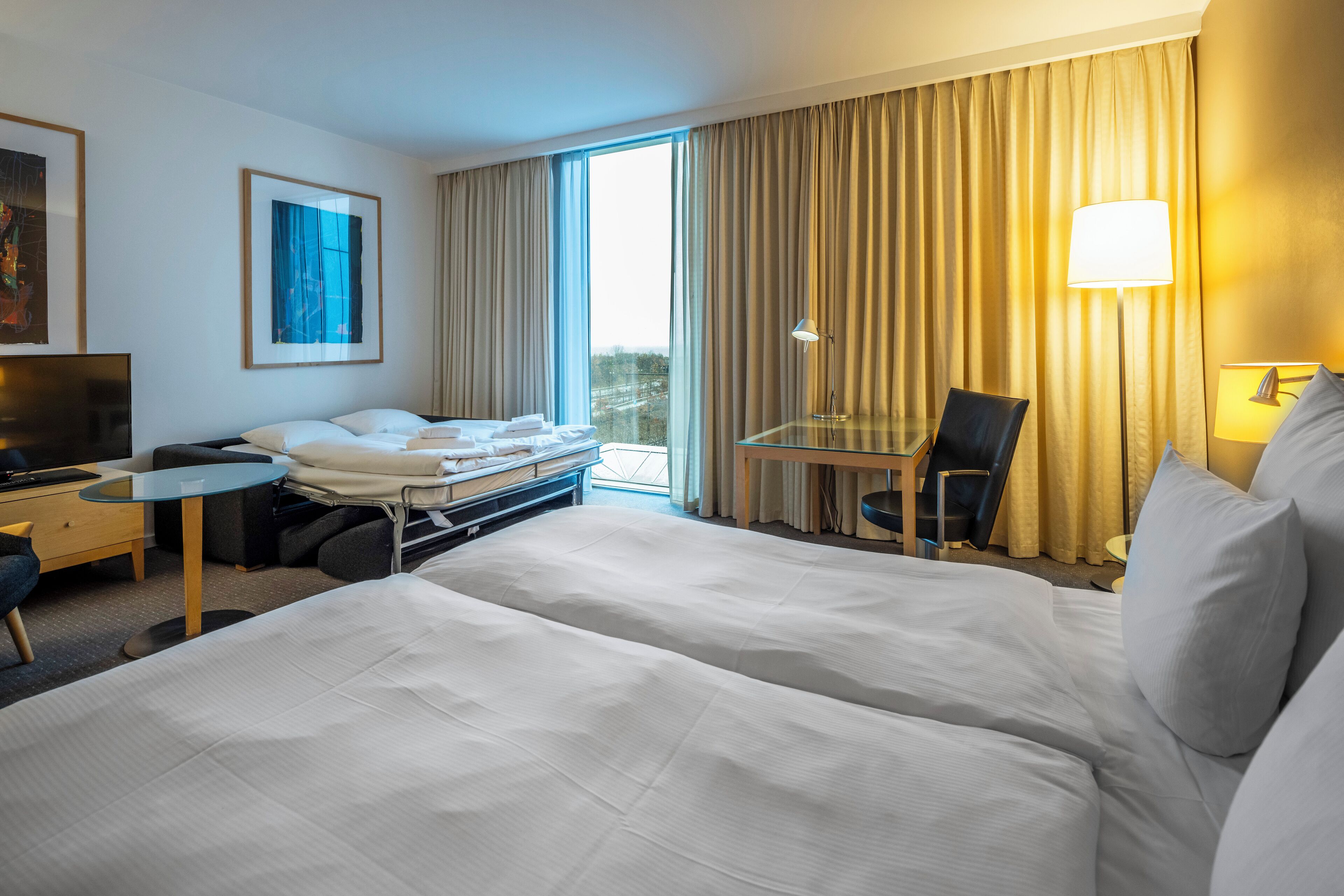 A hotel room with two beds, a desk, a chair, a TV, and a window with curtains.