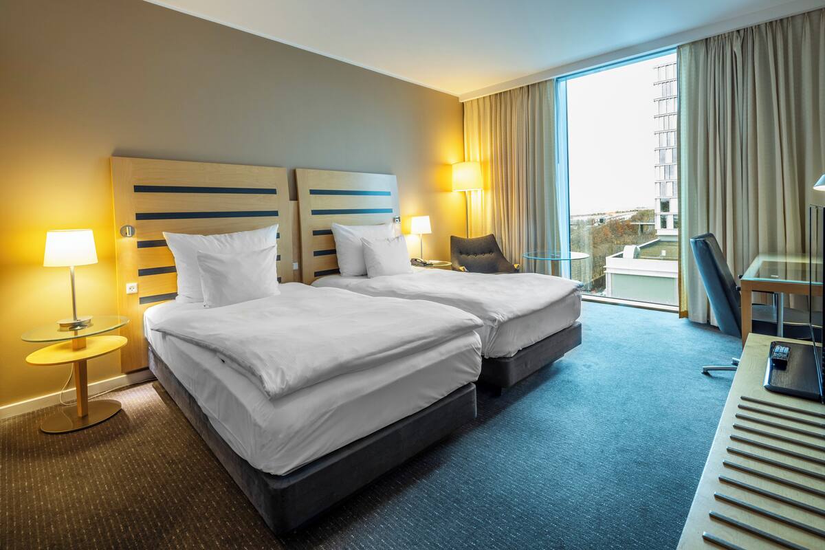 Ein Hotelzimmer mit zwei Betten, einem Schreibtisch, einem Stuhl und Blick auf die Stadt.