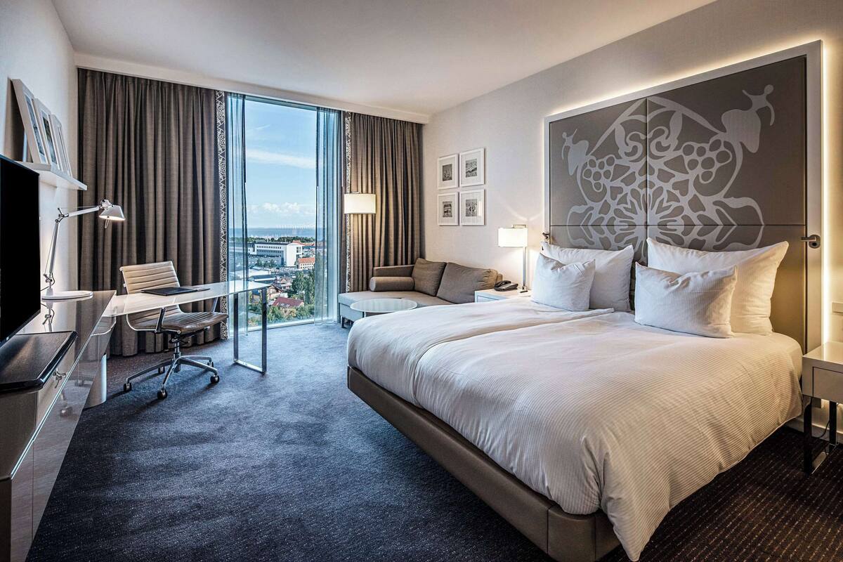 Ein modernes Hotelzimmer mit einem großen Bett, einem Schreibtisch, einem Sessel und Blick auf die Stadt durch große Fenster.