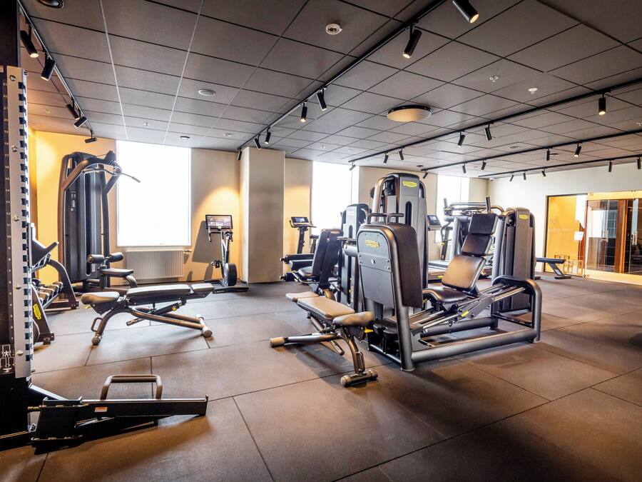 Uma academia com diversos equipamentos de exercício, uma janela grande e uma porta que dá para outro espaço.
