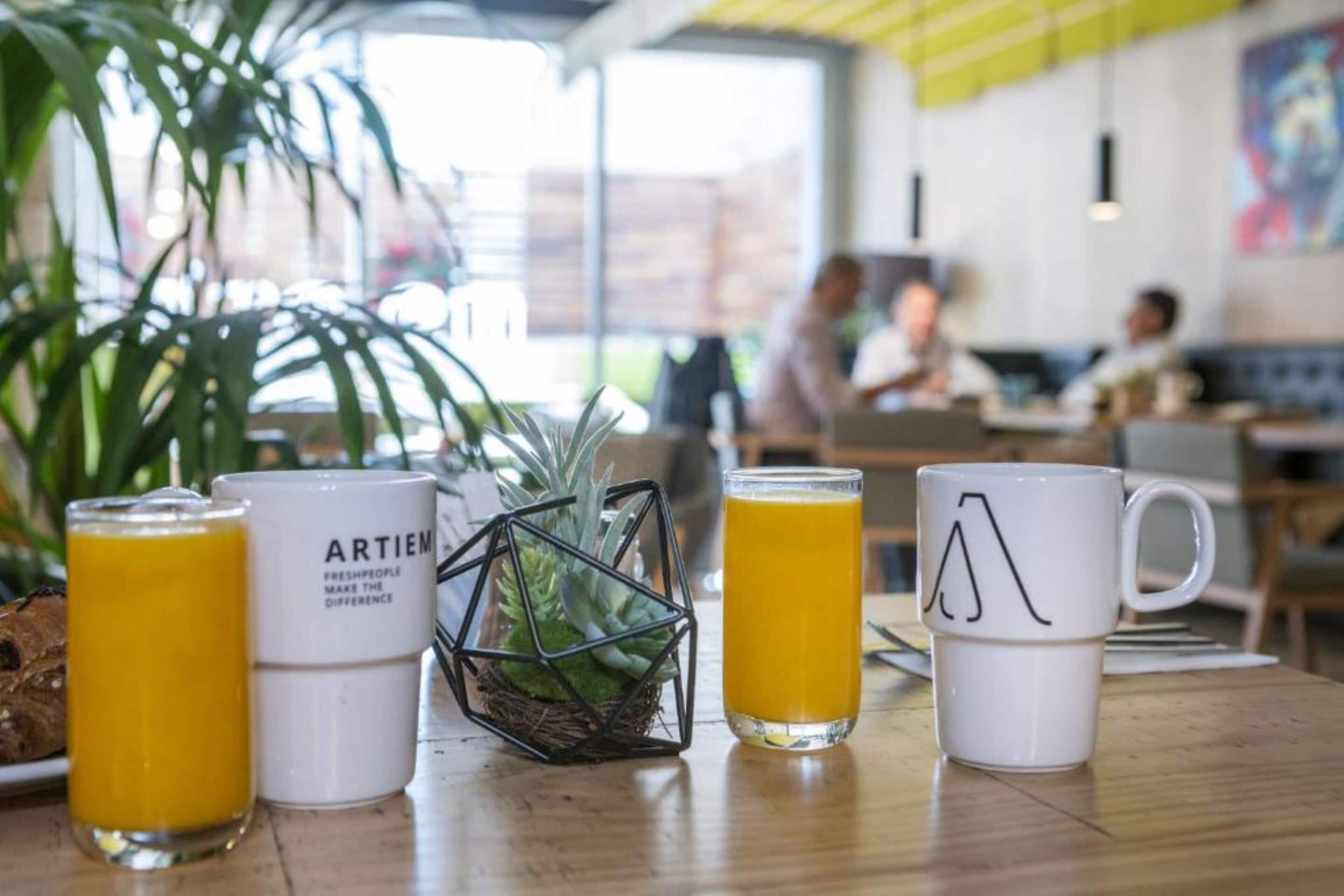 Una mesa con un vaso de jugo de naranja, una taza blanca con diseño geométrico y otra taza blanca con texto.