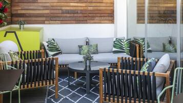 Un área de estar moderna al aire libre con un sofá, sillas y una mesa redonda, frente a una pared de madera y un panel de vidrio.