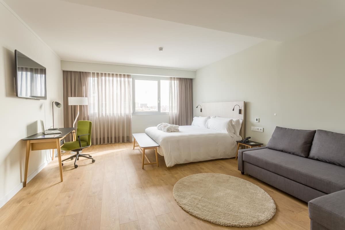 A modern hotel room with a bed, a desk, a green office chair, a grey sofa, and a round rug.