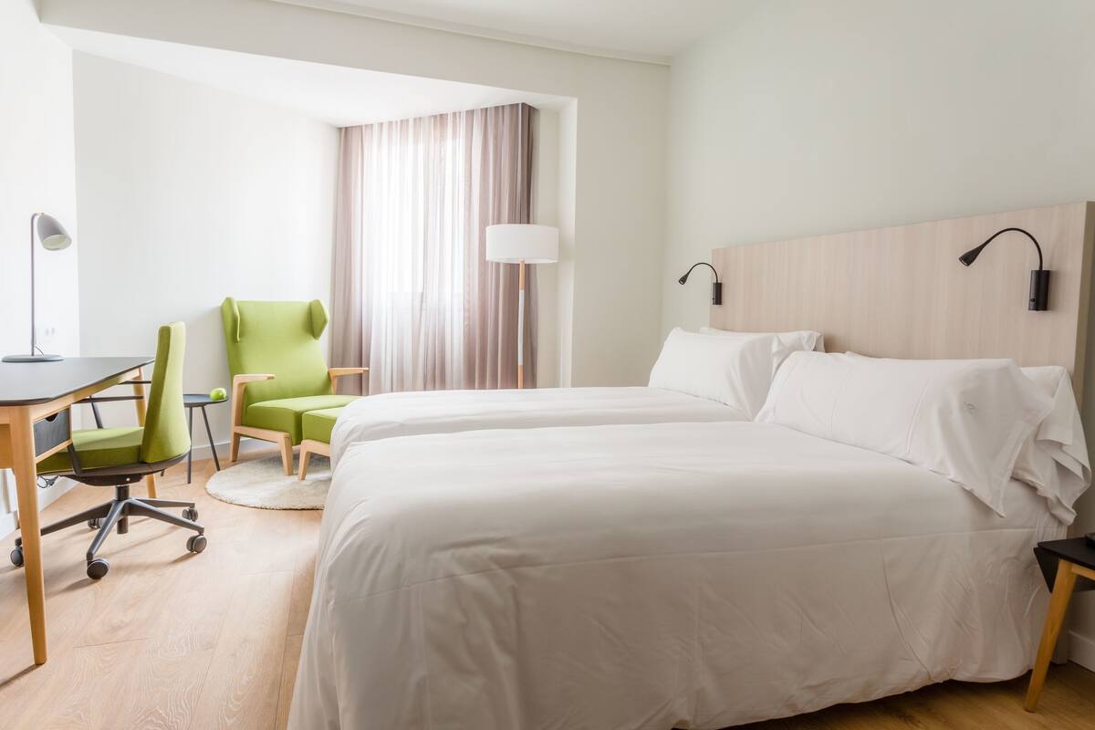 A hotel room with two beds, a desk with a chair, a lamp, and a window with curtains.