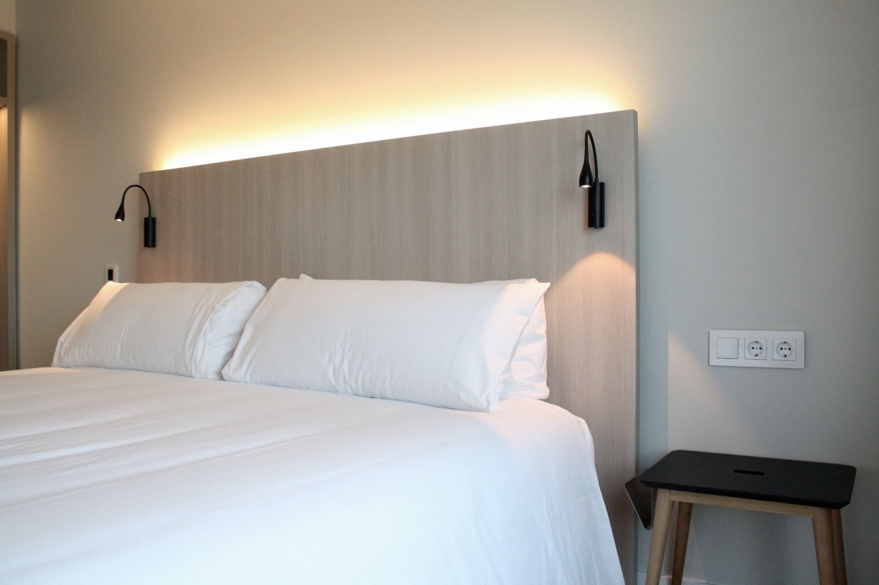 A neatly made bed with white linens, a small bedside table with a lamp, and a wall socket.