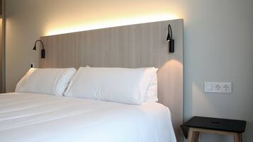 A neatly made bed with white linens, a small bedside table with a lamp, and a wall socket.