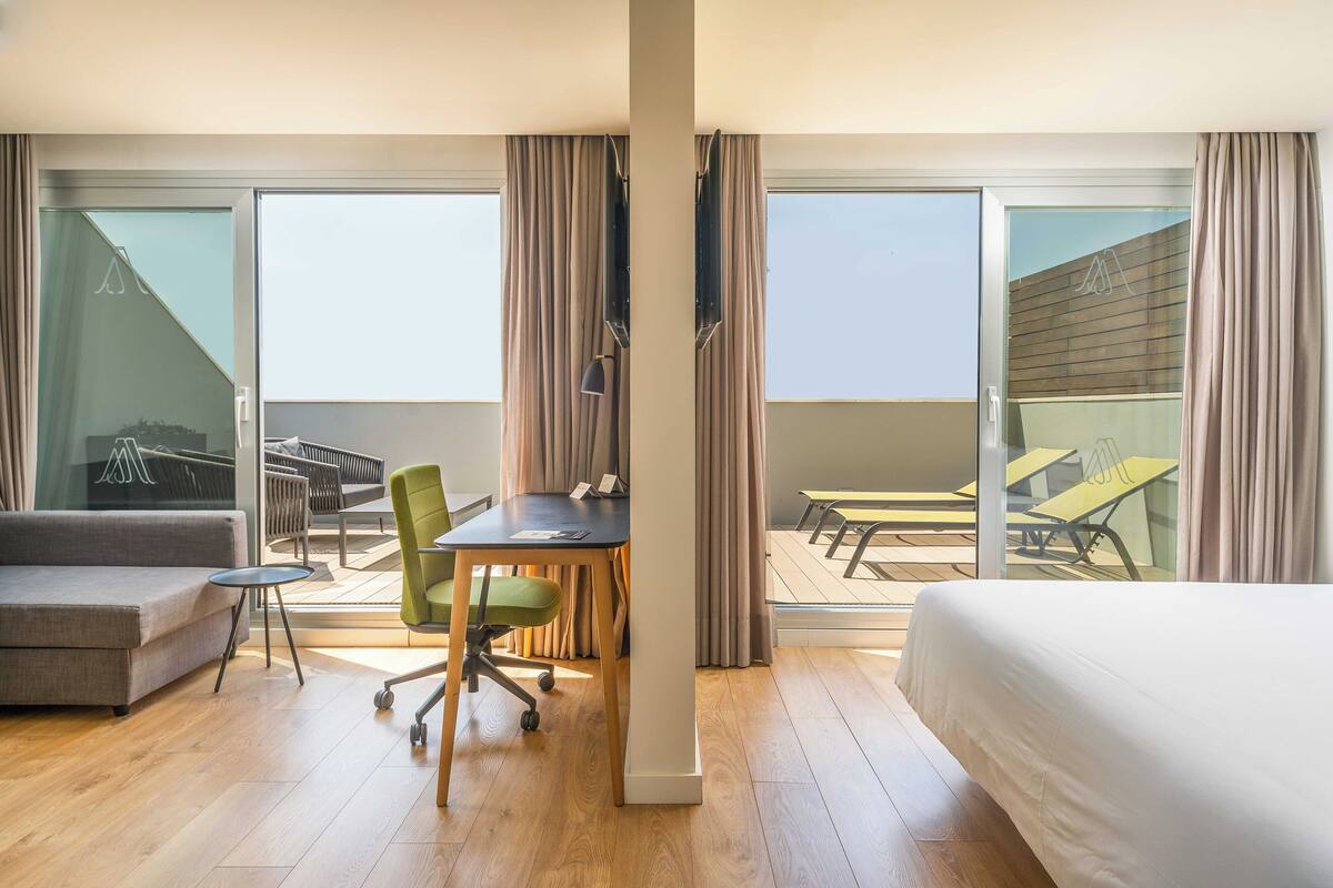 A hotel room with a bed, a desk, a chair, and a balcony with outdoor furniture.