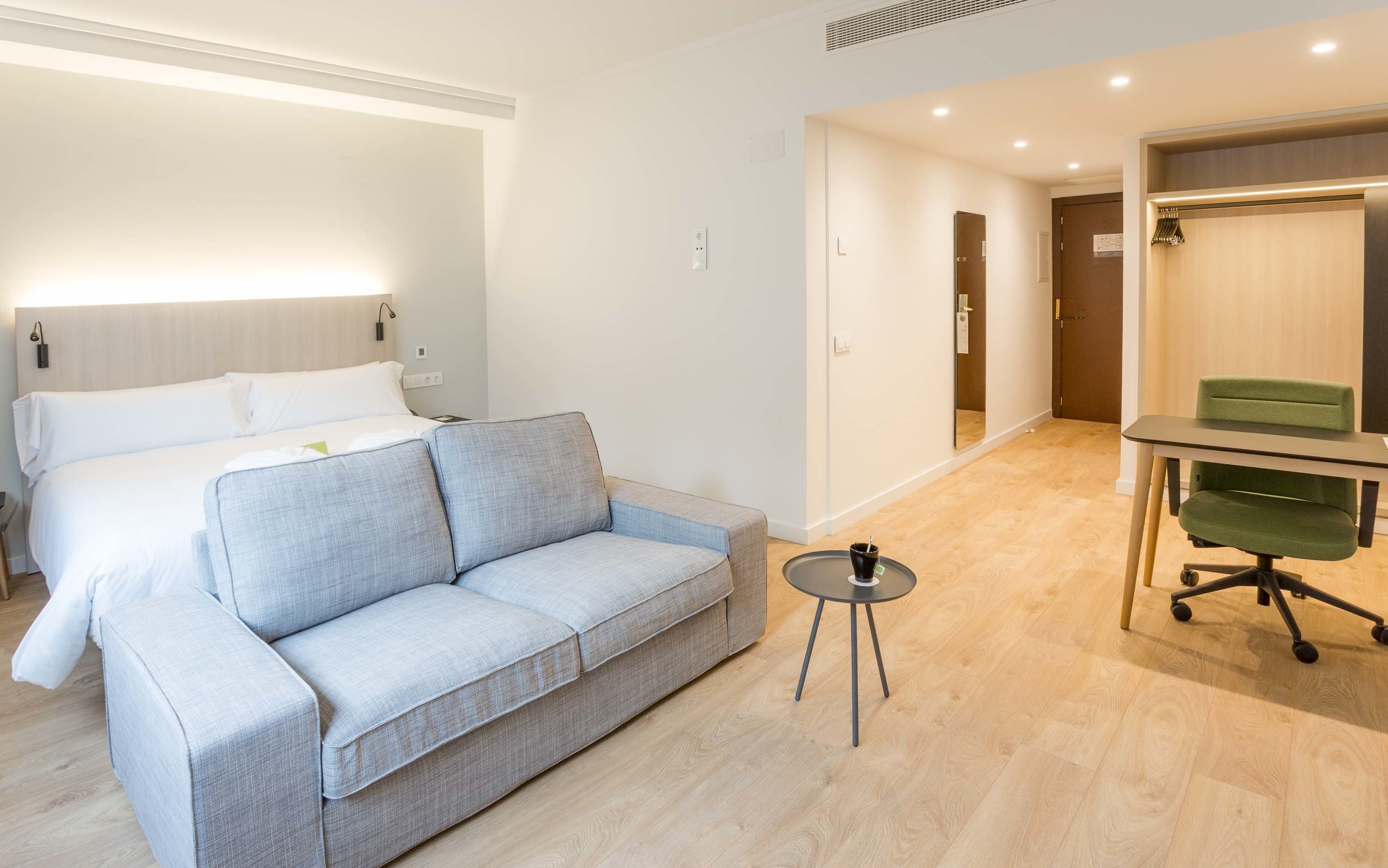Una camera d'albergo moderna con un letto, un divano grigio, un piccolo tavolo rotondo e una sedia da ufficio verde.