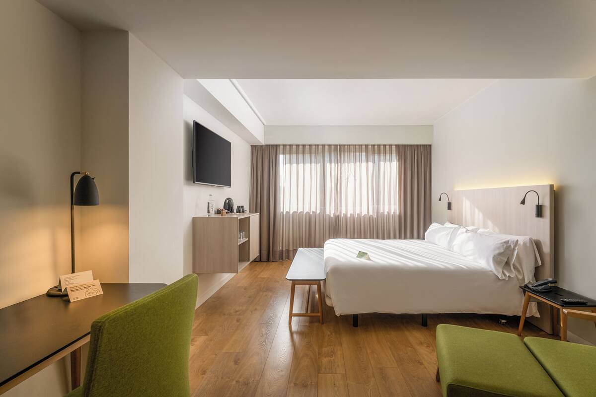 A hotel room with a bed, a desk, a chair, a small table, a TV, and a window with curtains.