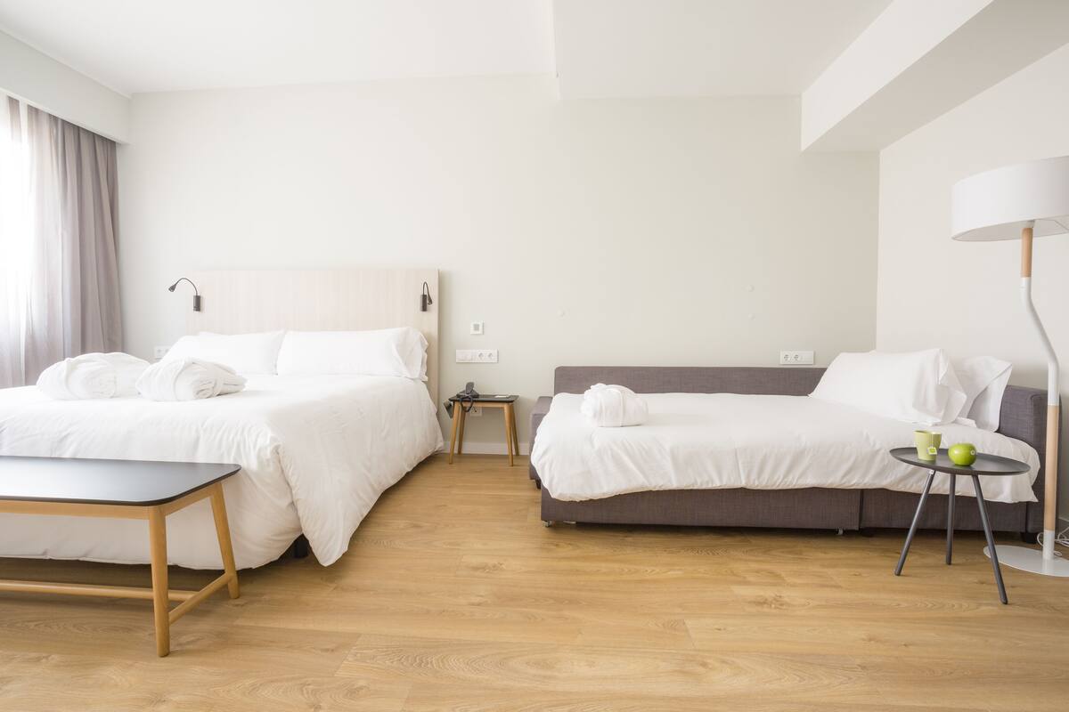 A hotel room with two beds, a small table, a sofa, a lamp, and a bedside table with a green cup and apple.