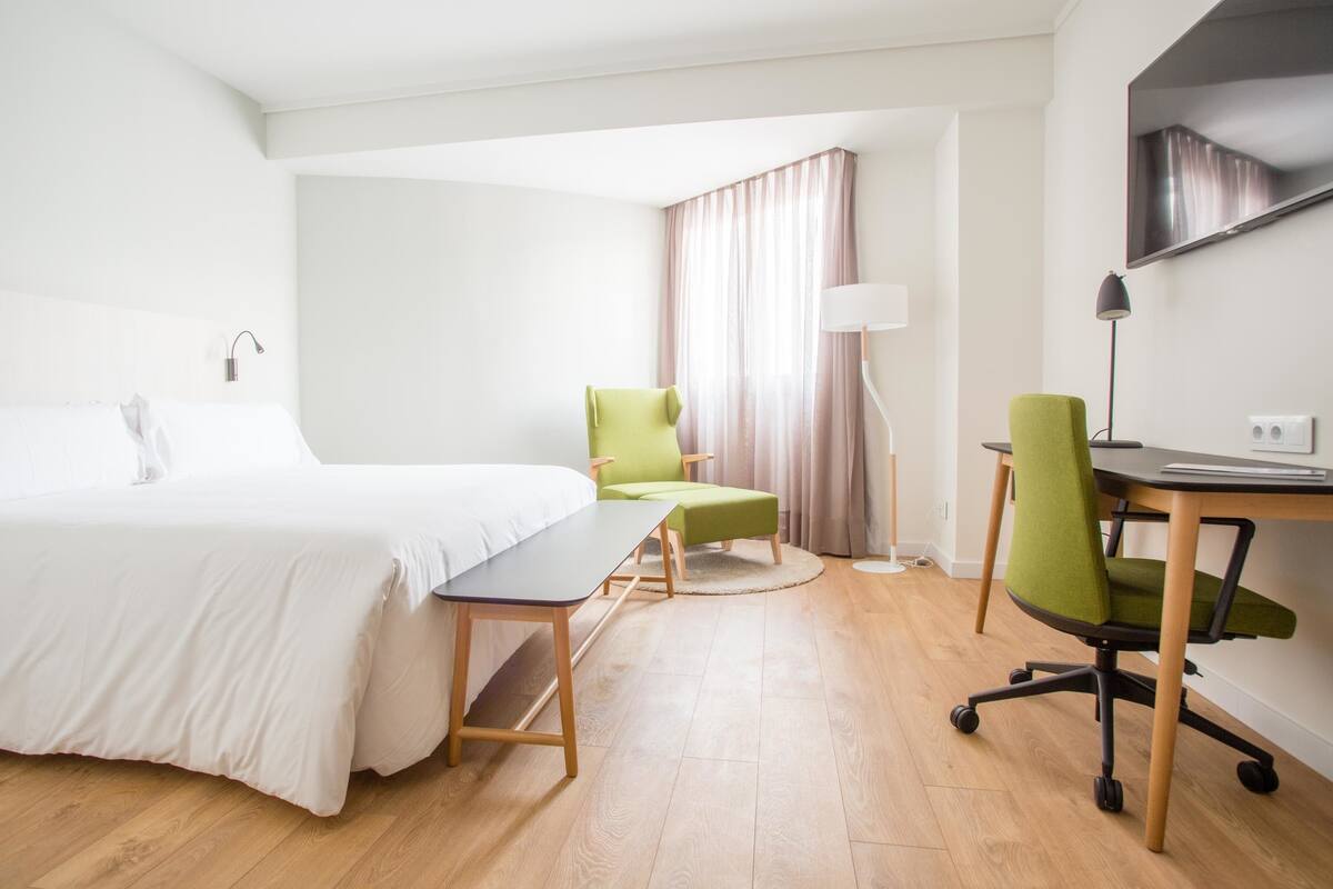 A hotel room with a bed, a desk, two chairs, a lamp, and a TV.