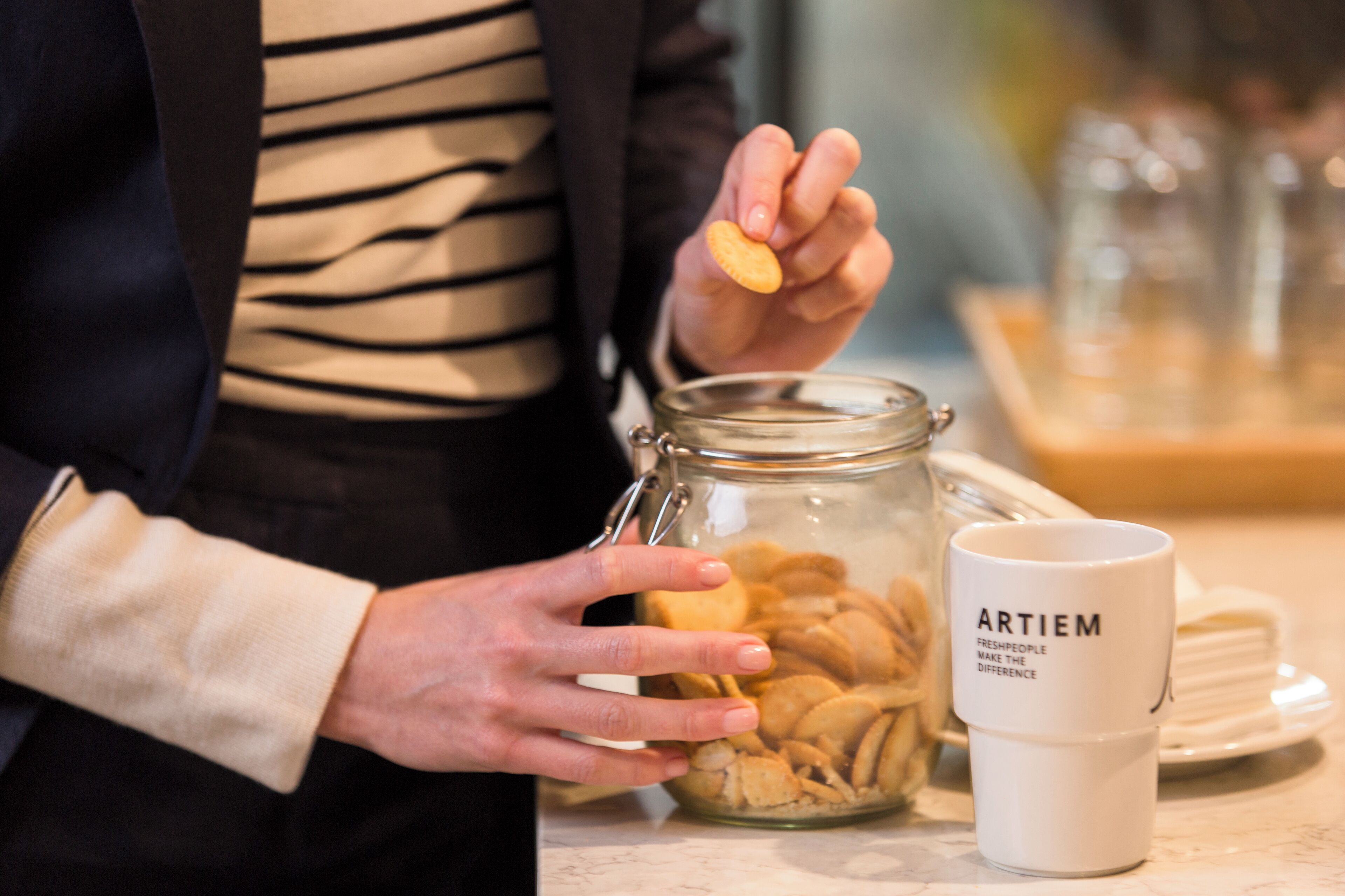 Una persona sosteniendo una galleta de un tarro, con una taza que dice 'ARTIEM' sobre una mesa.