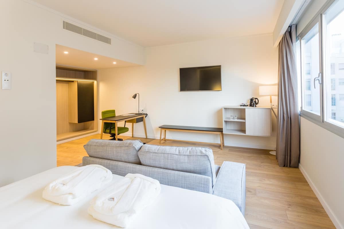 A modern hotel room with a bed, a sofa, a desk, and a TV on the wall.