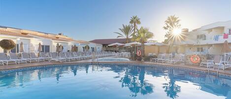 A clear swimming pool with sunlit surroundings, lounge chairs, and palm trees.