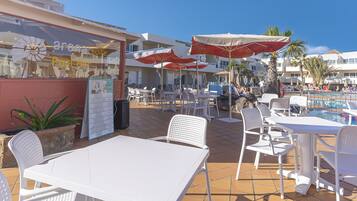 A poolside area with white tables and chairs, red and white umbrellas, and a pool in the background.
