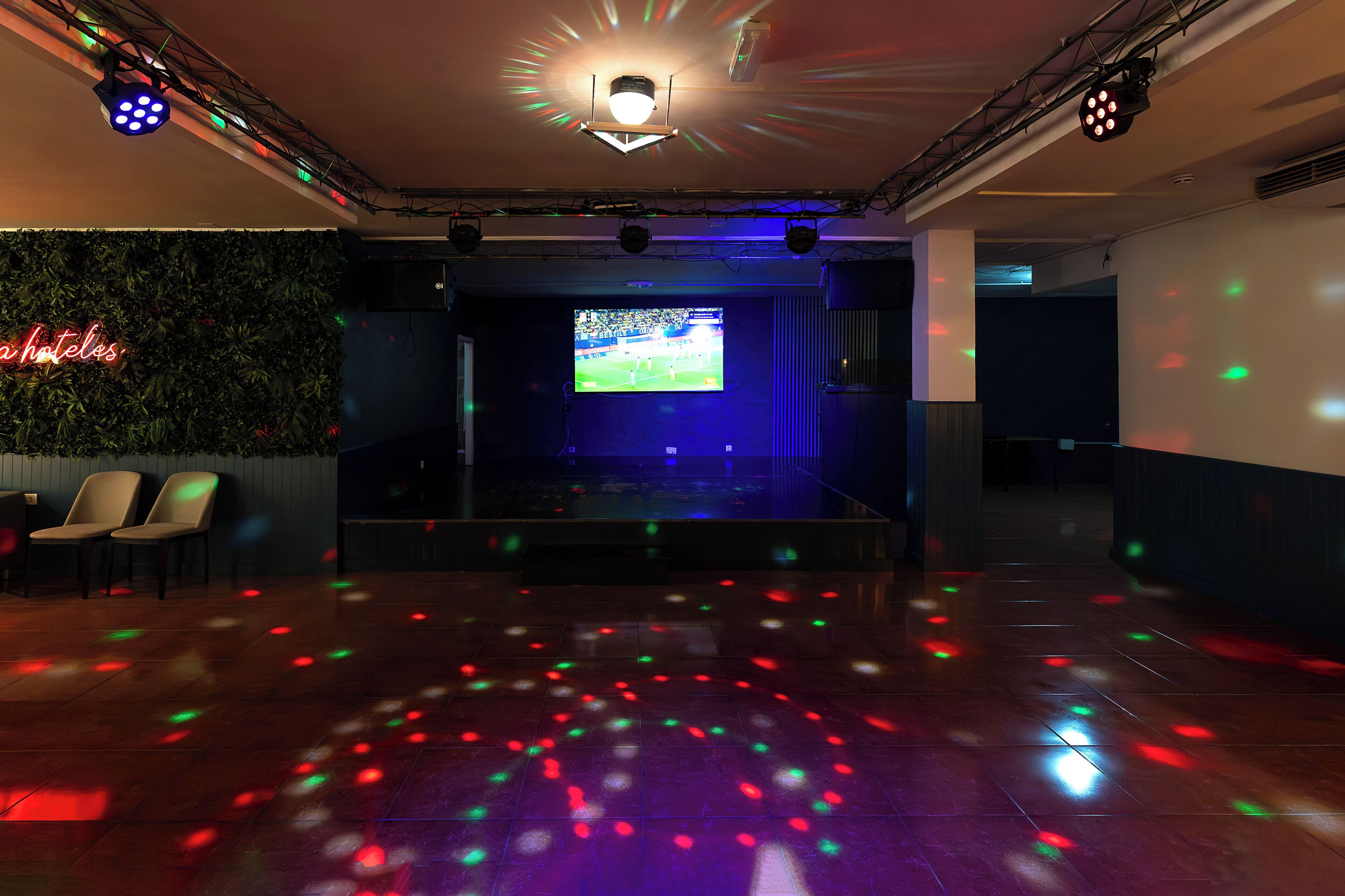 A room with a stage, chairs, a TV screen, and a wall with a neon sign.
