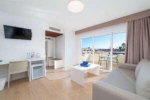 A modern hotel room with a flat-screen TV, a desk with a chair, a small table with a towel, a sofa, and a sliding glass door leading to a balcony with a view.
