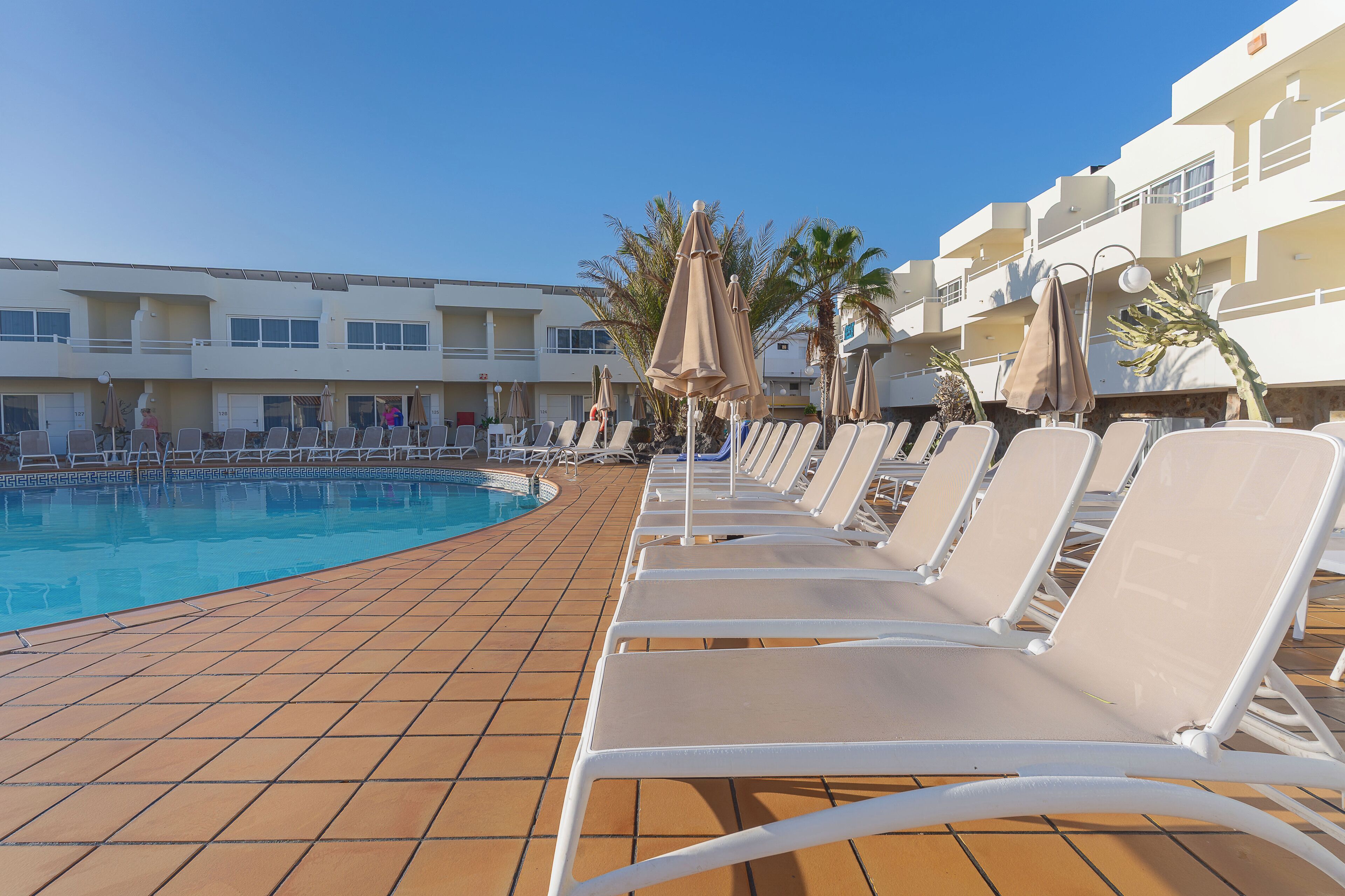 A poolside area with lounge chairs and umbrellas, a tiled deck, and a building with balconies in the background.