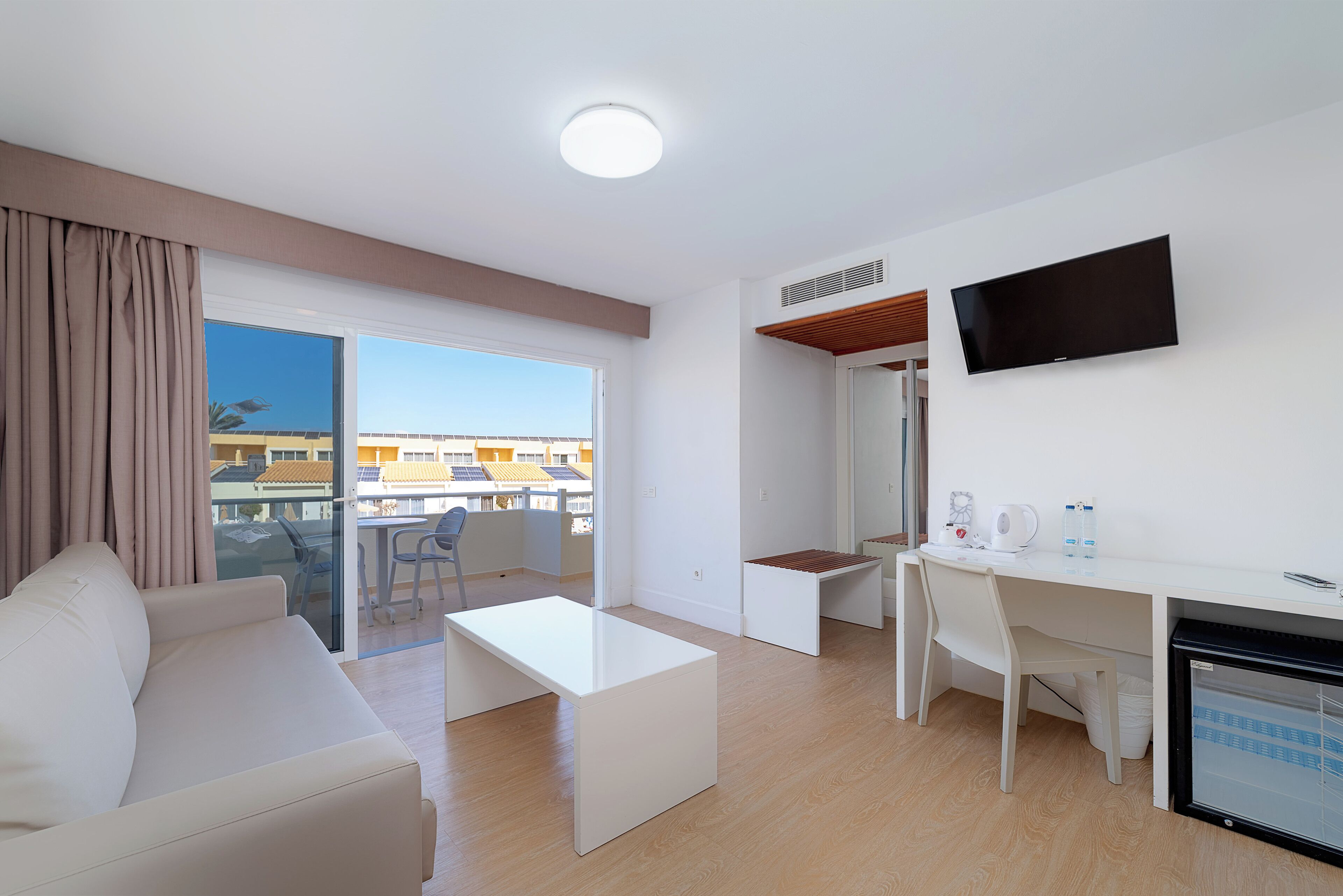 Una camera d'albergo moderna con un divano bianco, un piccolo tavolino, una scrivania con una sedia, una TV a schermo piatto e un mini-frigorifero.