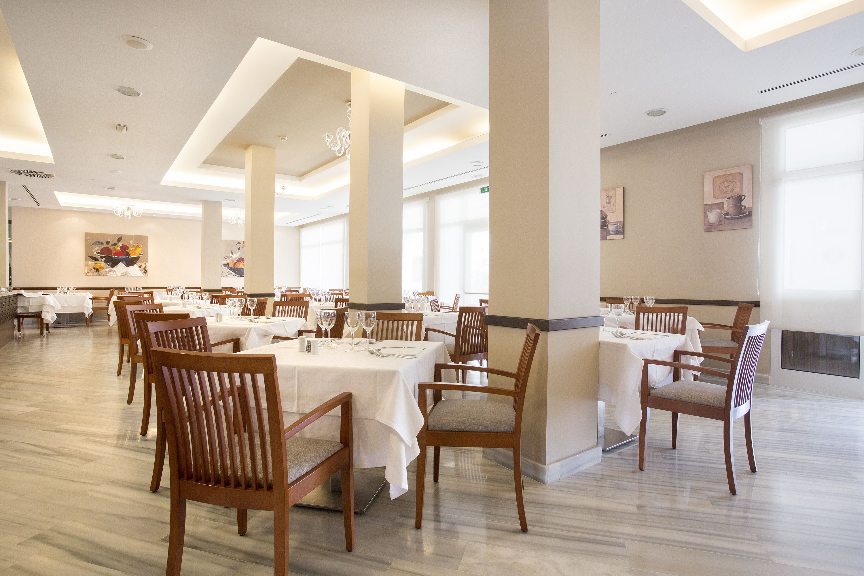 Uma ampla sala de jantar com várias mesas e cadeiras, tetos altos com iluminação embutida e grandes janelas que permitem a entrada de luz natural.