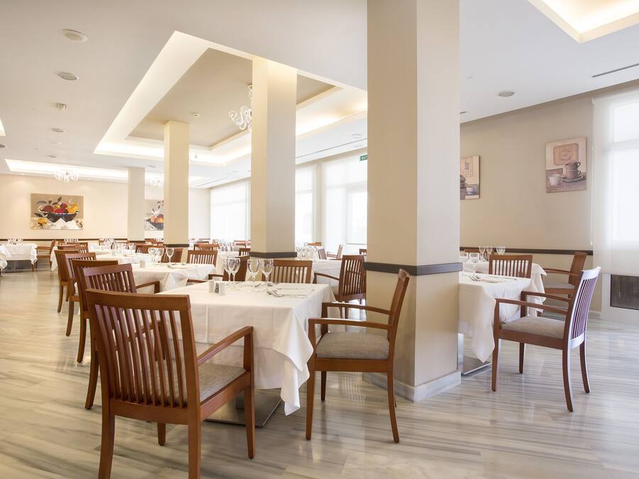 Uma ampla sala de jantar com várias mesas e cadeiras, tetos altos com iluminação embutida e grandes janelas que permitem a entrada de luz natural.
